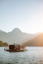 Floating sauna with people on ocean surrounded by mountains at sunset.