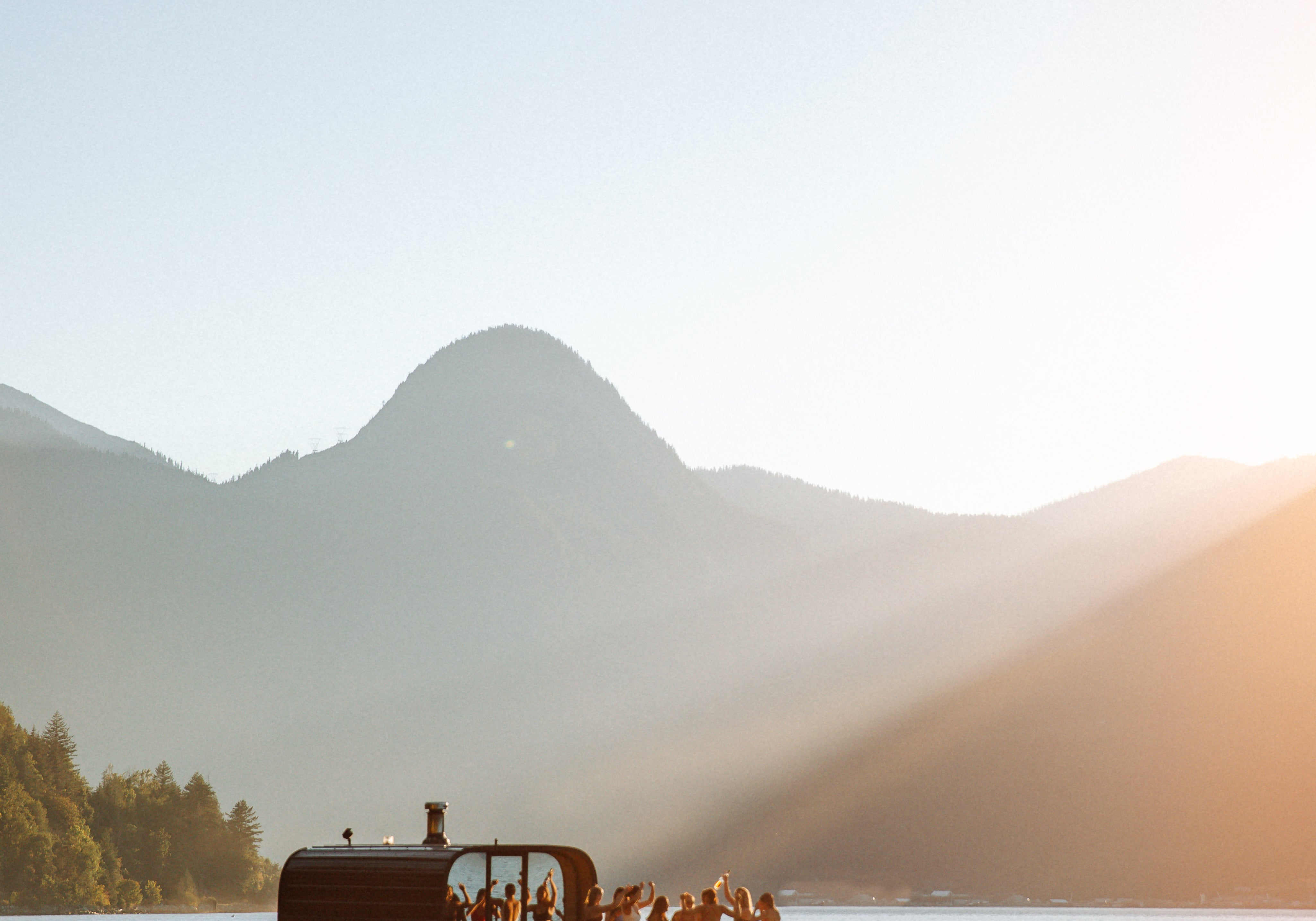 Floating sauna with people on ocean surrounded by mountains at sunset.