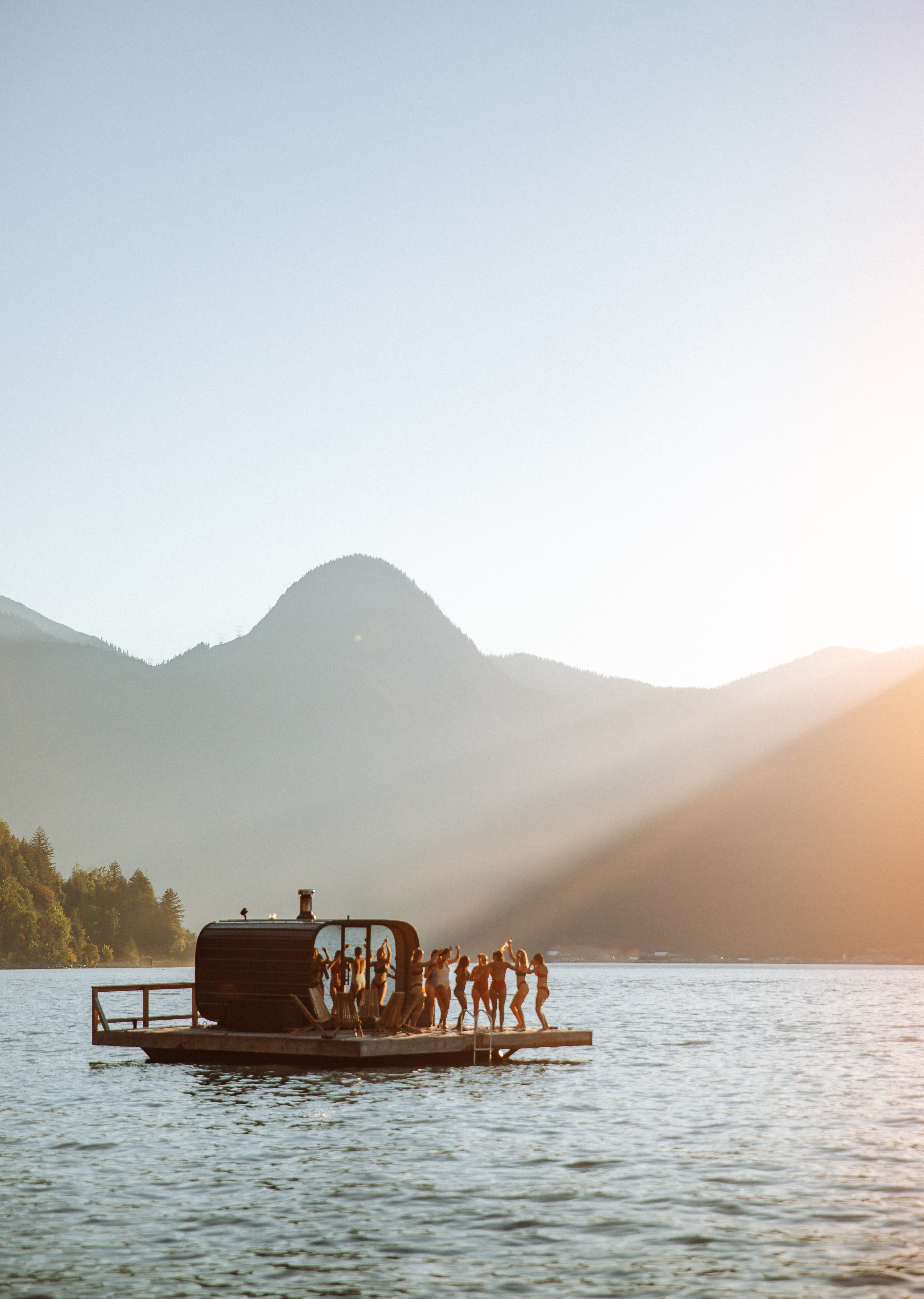 Floating sauna with people on ocean surrounded by mountains at sunset.